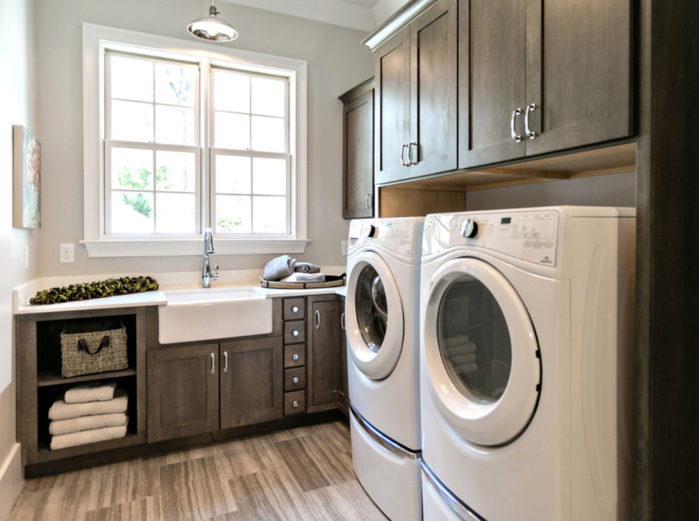 Small transitional l-shaped dedicated laundry room in Bridgeport with a farmhouse sink, shaker cabinets, dark wood cabinets, quartz benchtops, grey walls, porcelain floors, a side-by-side washer and dryer, brown floor and white benchtop.
