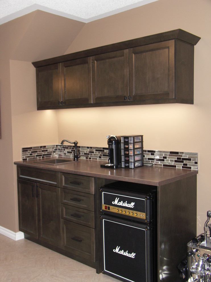 This is an example of a small transitional single-wall wet bar in Edmonton with recessed-panel cabinets, laminate benchtops, multi-coloured splashback, a drop-in sink, dark wood cabinets, ceramic floors, beige floor and glass tile splashback.