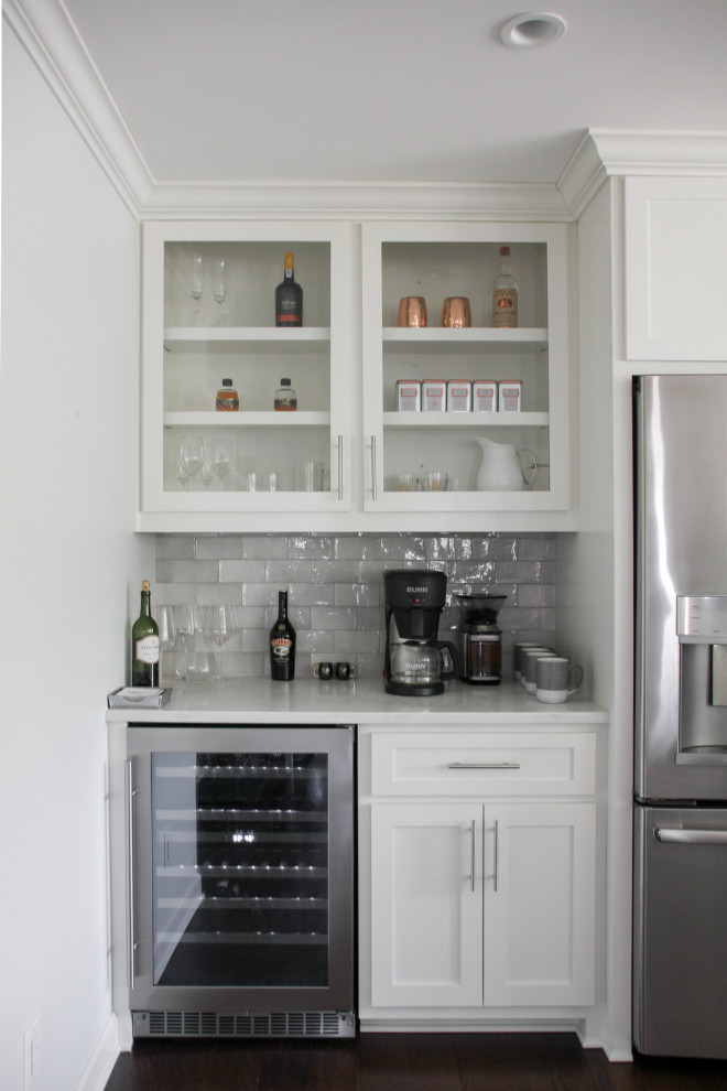 This is an example of a small beach style l-shaped home bar in Charlotte with no sink, shaker cabinets, white cabinets, quartz benchtops, grey splashback, ceramic splashback, dark hardwood floors, brown floor and white benchtop.
