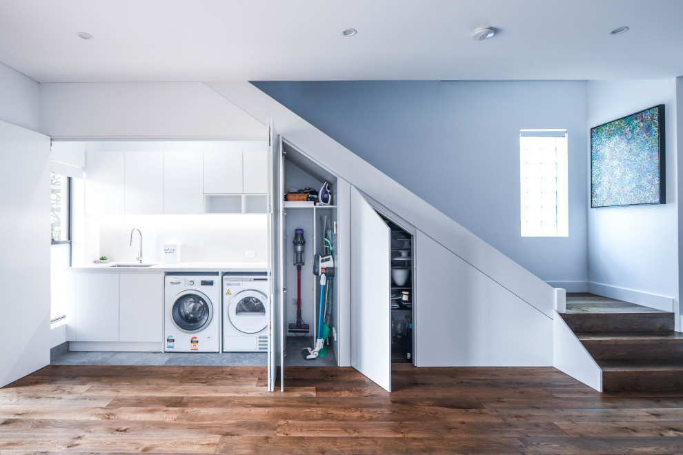 Mid-sized modern single-wall dedicated laundry room in Sydney with an undermount sink, flat-panel cabinets, white cabinets, quartz benchtops, white splashback, ceramic splashback, white walls, medium hardwood floors, a side-by-side washer and dryer, brown floor and white benchtop.