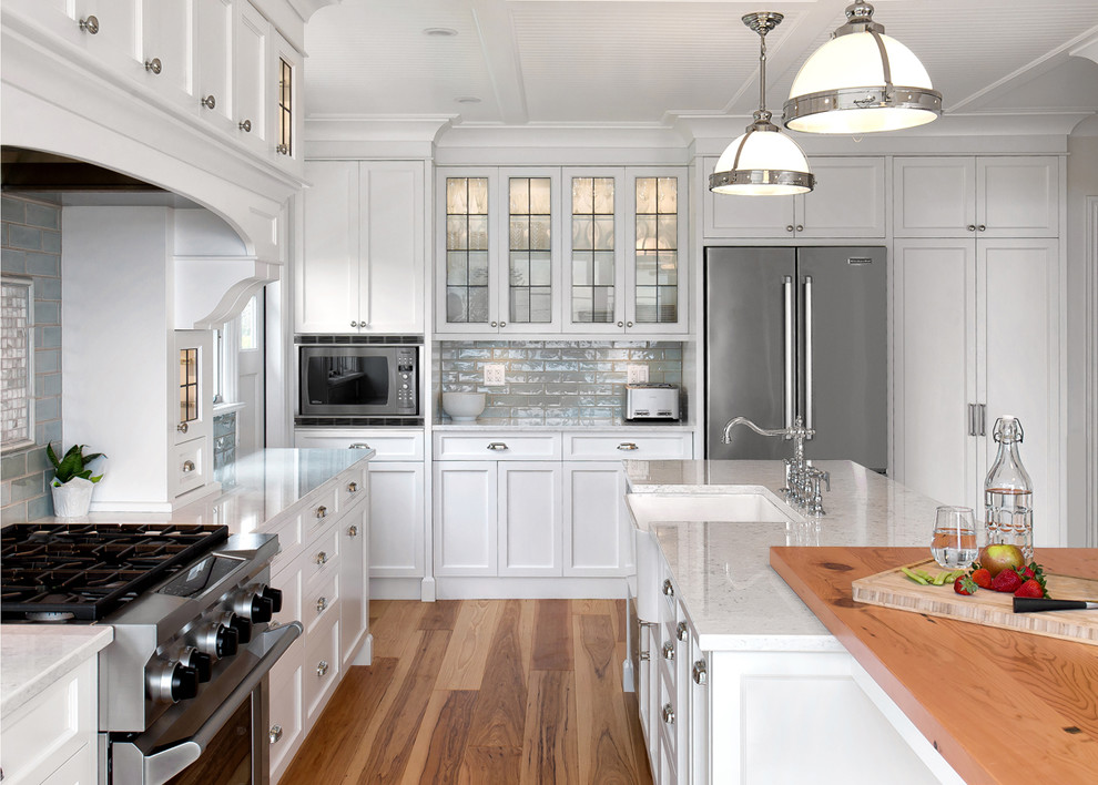 This is an example of a beach style u-shaped kitchen in Vancouver with a farmhouse sink, recessed-panel cabinets, white cabinets, grey splashback, stainless steel appliances, medium hardwood floors and with island.