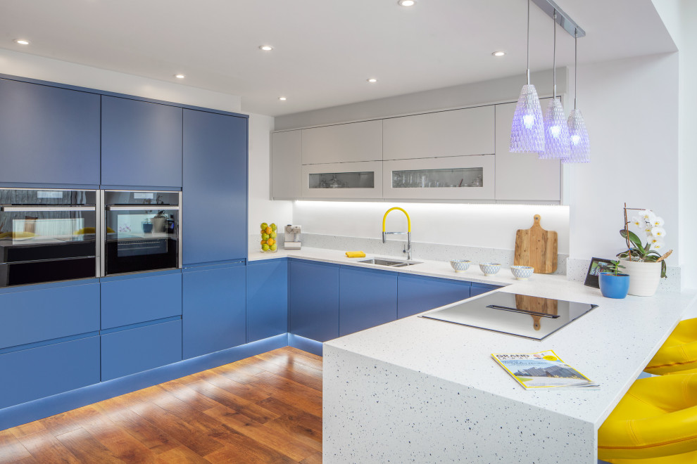 This is an example of a medium sized contemporary u-shaped kitchen/diner in Other with a submerged sink, flat-panel cabinets, blue cabinets, composite countertops, integrated appliances, dark hardwood flooring, a breakfast bar, white worktops and feature lighting.