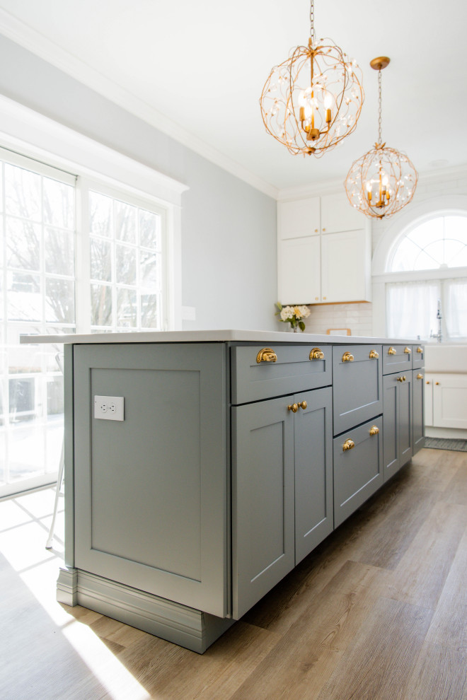 This is an example of a large l-shaped enclosed kitchen in New York with a belfast sink, shaker cabinets, blue cabinets, engineered stone countertops, white splashback, metro tiled splashback, stainless steel appliances, light hardwood flooring, an island, brown floors and white worktops.