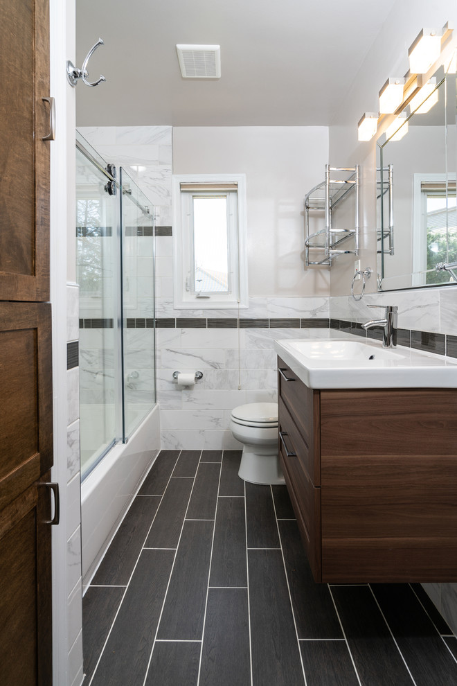 Photo of a mid-sized contemporary 3/4 bathroom in DC Metro with beaded inset cabinets, brown cabinets, a shower/bathtub combo, a one-piece toilet, an integrated sink, marble benchtops, a sliding shower screen, gray tile, marble, grey walls, vinyl floors, black floor, white benchtops and an alcove tub.