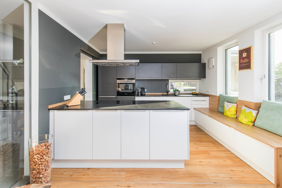 Inspiration for a mid-sized contemporary galley eat-in kitchen in Nuremberg with white cabinets, granite benchtops, flat-panel cabinets, black splashback, panelled appliances, light hardwood floors, a peninsula, beige floor and black benchtop.