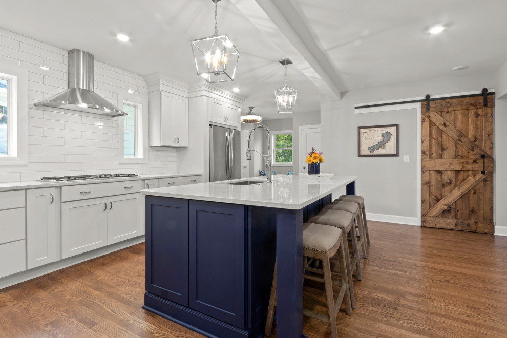 This is an example of an expansive modern l-shaped open plan kitchen in Milwaukee with an undermount sink, shaker cabinets, white cabinets, quartz benchtops, white splashback, cement tile splashback, stainless steel appliances, medium hardwood floors, with island, brown floor and white benchtop.