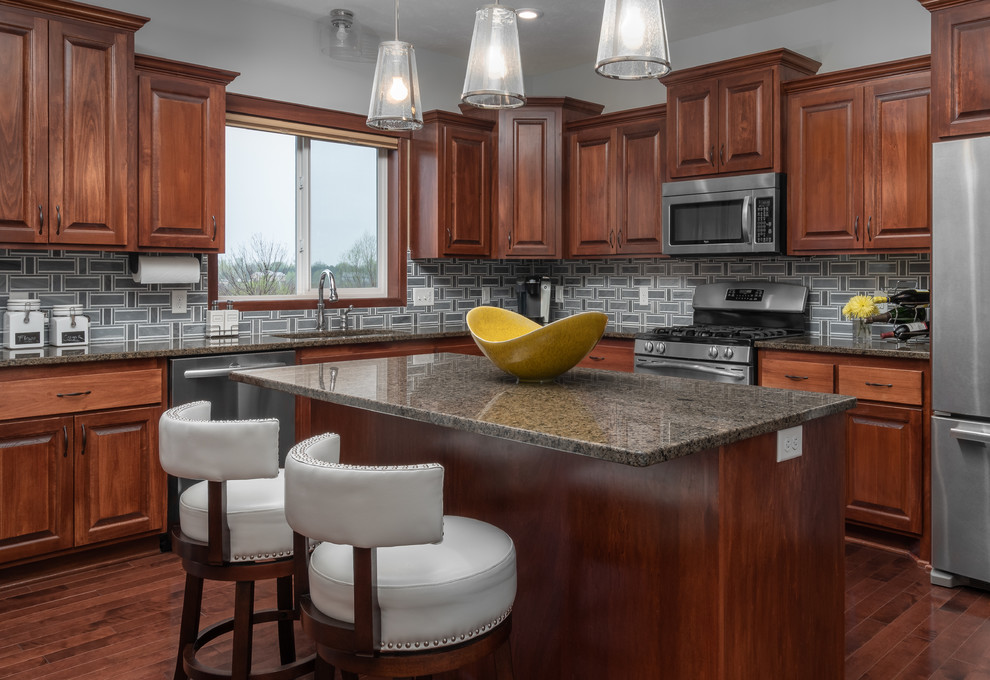 Traditional l-shaped kitchen in Minneapolis with raised-panel cabinets, dark wood cabinets, grey splashback, stainless steel appliances, dark hardwood floors, with island, brown floor, brown benchtop and granite benchtops.