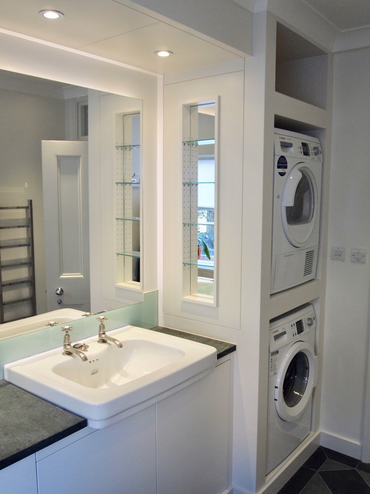 Photo of a mid-sized contemporary galley utility room in London with a drop-in sink, flat-panel cabinets, white cabinets, granite benchtops, white walls, slate floors, a stacked washer and dryer, green floor and grey benchtop.