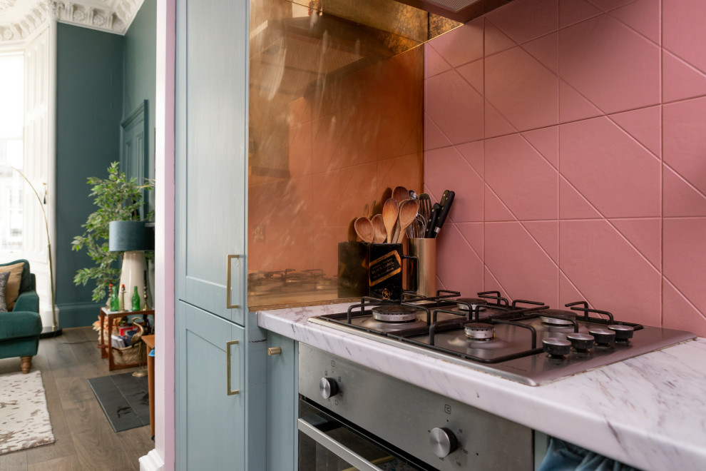 Small eclectic l-shaped open plan kitchen in Edinburgh with a single-bowl sink, shaker cabinets, blue cabinets, laminate benchtops, pink splashback, ceramic splashback, stainless steel appliances, vinyl floors, no island, multi-coloured floor and grey benchtop.