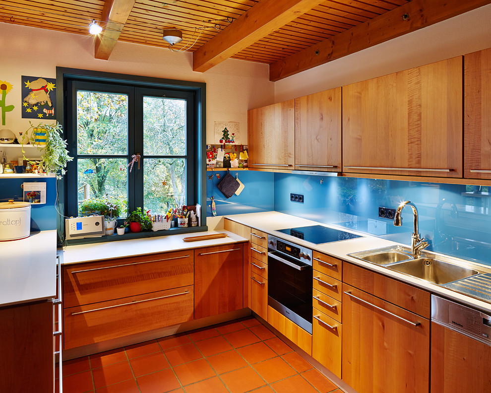 Country open plan kitchen in Dusseldorf with a drop-in sink, flat-panel cabinets, medium wood cabinets, glass benchtops, blue splashback, glass sheet splashback, coloured appliances, terra-cotta floors and a peninsula.