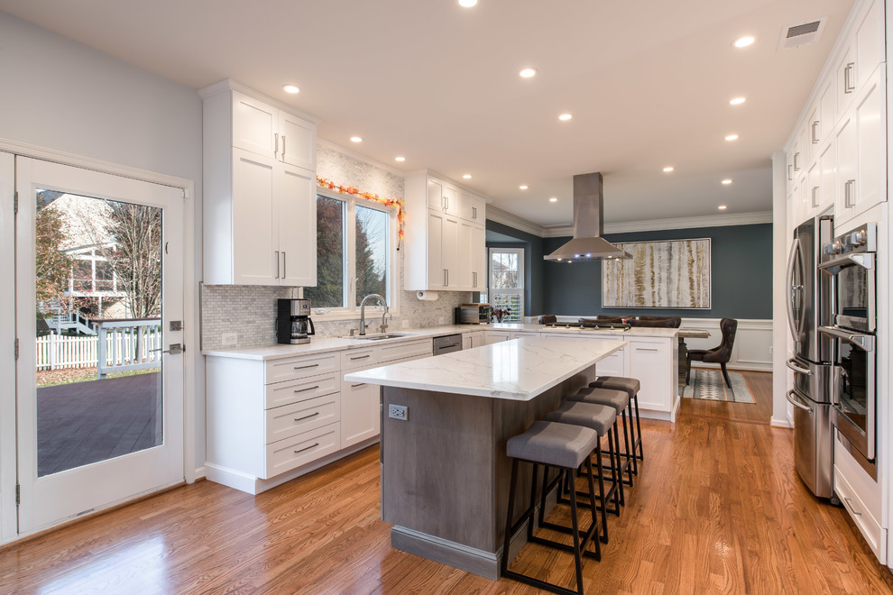 This is an example of a transitional u-shaped eat-in kitchen in DC Metro with an undermount sink, shaker cabinets, white cabinets, grey splashback, stainless steel appliances, medium hardwood floors, with island and brown floor.