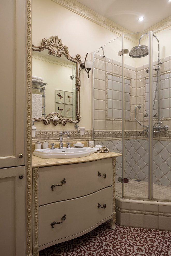 This is an example of a small traditional 3/4 bathroom in Moscow with beige tile, ceramic tile, ceramic floors, marble benchtops, red floor, flat-panel cabinets, beige cabinets, a corner shower, beige walls, a drop-in sink, a hinged shower door and beige benchtops.