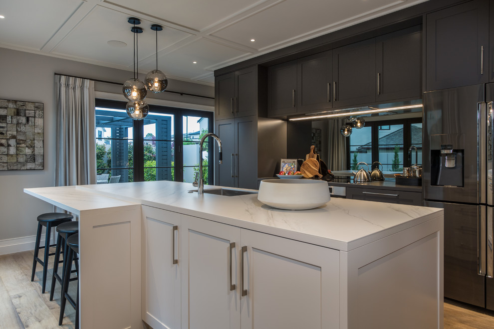 Large traditional eat-in kitchen in Auckland with white cabinets, mirror splashback, stainless steel appliances, medium hardwood floors, with island, a drop-in sink, recessed-panel cabinets, tile benchtops, metallic splashback and brown floor.