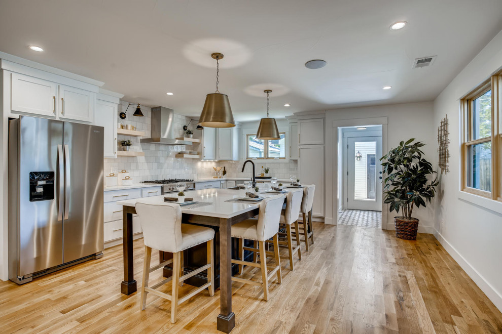 Example of a mid-sized transitional l-shaped eat-in kitchen design in Denver with a farmhouse sink, shaker cabinets, white cabinets, quartzite countertops, white backsplash, porcelain backsplash, stainless steel appliances and an island