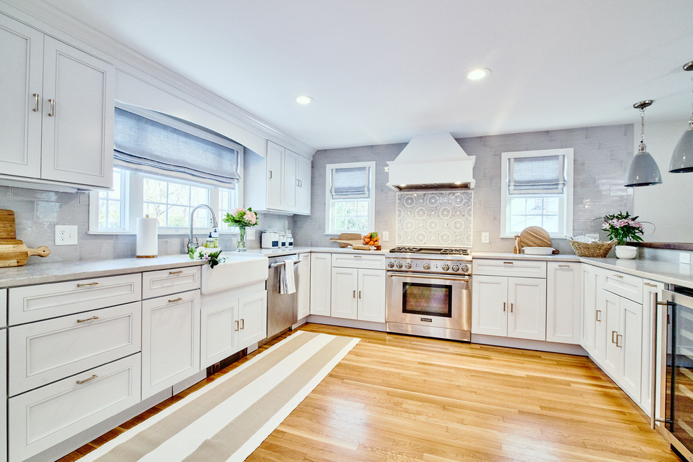Photo of a large transitional u-shaped open plan kitchen in Providence with a farmhouse sink, shaker cabinets, white cabinets, quartz benchtops, grey splashback, mosaic tile splashback, stainless steel appliances, light hardwood floors, with island, beige floor and white benchtop.
