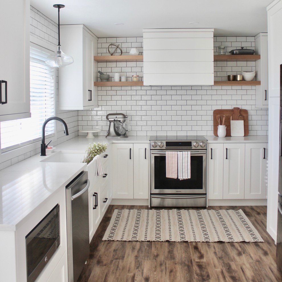 Mid-sized country l-shaped separate kitchen in Vancouver with a farmhouse sink, shaker cabinets, white cabinets, solid surface benchtops, white splashback, ceramic splashback, stainless steel appliances, laminate floors, no island, brown floor and white benchtop.