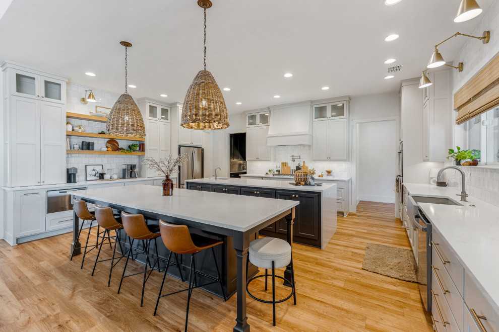 Example of a mid-sized transitional u-shaped light wood floor kitchen design in Dallas with a farmhouse sink, shaker cabinets, gray cabinets, quartz countertops, white backsplash, porcelain backsplash, stainless steel appliances, two islands and white countertops