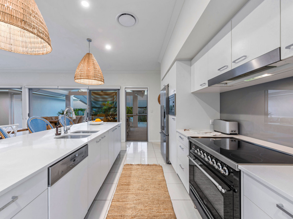 Photo of a mid-sized beach style galley eat-in kitchen in Brisbane with an undermount sink, flat-panel cabinets, white cabinets, quartz benchtops, grey splashback, glass sheet splashback, black appliances, ceramic floors, with island, white floor and white benchtop.