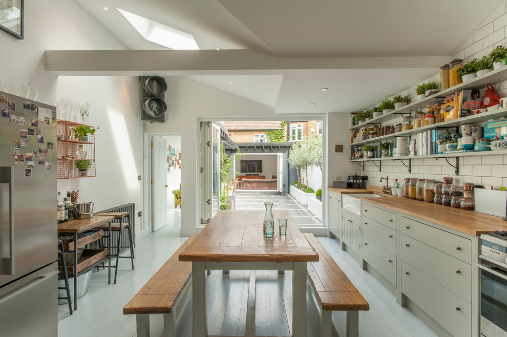 This is an example of a country kitchen in London with a farmhouse sink, flat-panel cabinets, green cabinets, white splashback, subway tile splashback, stainless steel appliances and light hardwood floors.