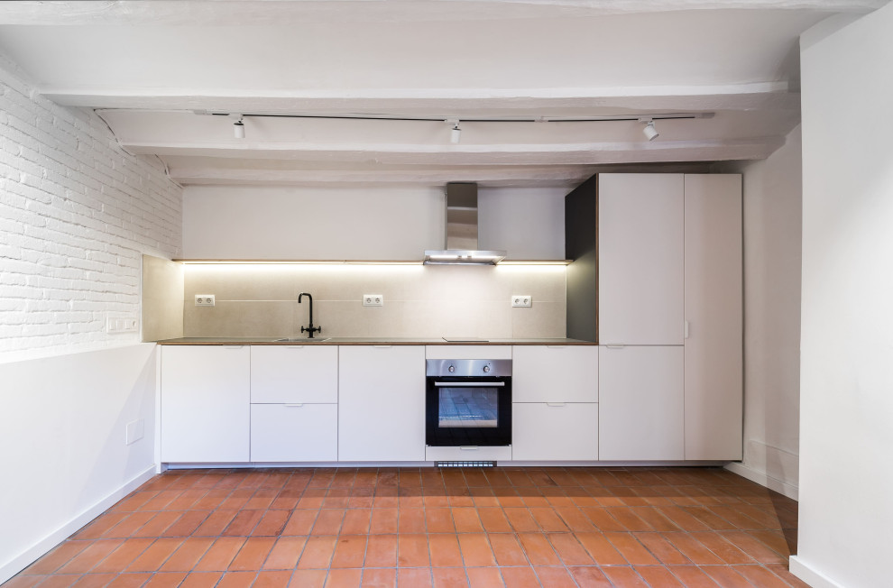 This is an example of a mid-sized transitional single-wall eat-in kitchen in Barcelona with a single-bowl sink, white cabinets, black splashback, stainless steel appliances, brick floors, orange floor, black benchtop and vaulted.