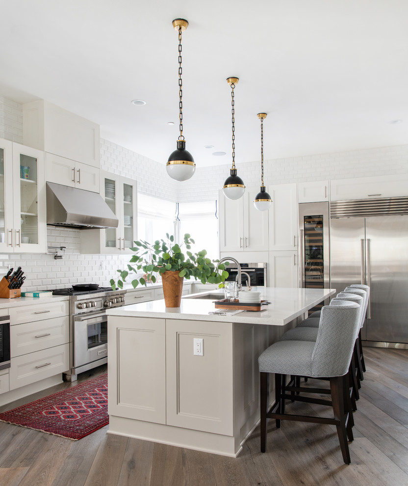 Design ideas for a transitional l-shaped kitchen in Los Angeles with an undermount sink, white cabinets, white splashback, subway tile splashback, stainless steel appliances, dark hardwood floors, with island, brown floor, white benchtop and glass-front cabinets.