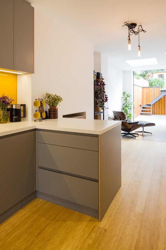 Inspiration for a mid-sized contemporary l-shaped open plan kitchen in London with an undermount sink, flat-panel cabinets, grey cabinets, solid surface benchtops, yellow splashback, glass sheet splashback, panelled appliances, bamboo floors, no island and brown floor.