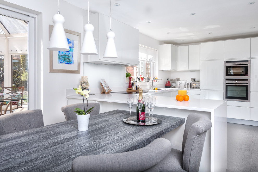 This is an example of a contemporary u-shaped kitchen/diner in Devon with flat-panel cabinets, stainless steel appliances, white cabinets, glass sheet splashback, white splashback, a built-in sink and quartz worktops.