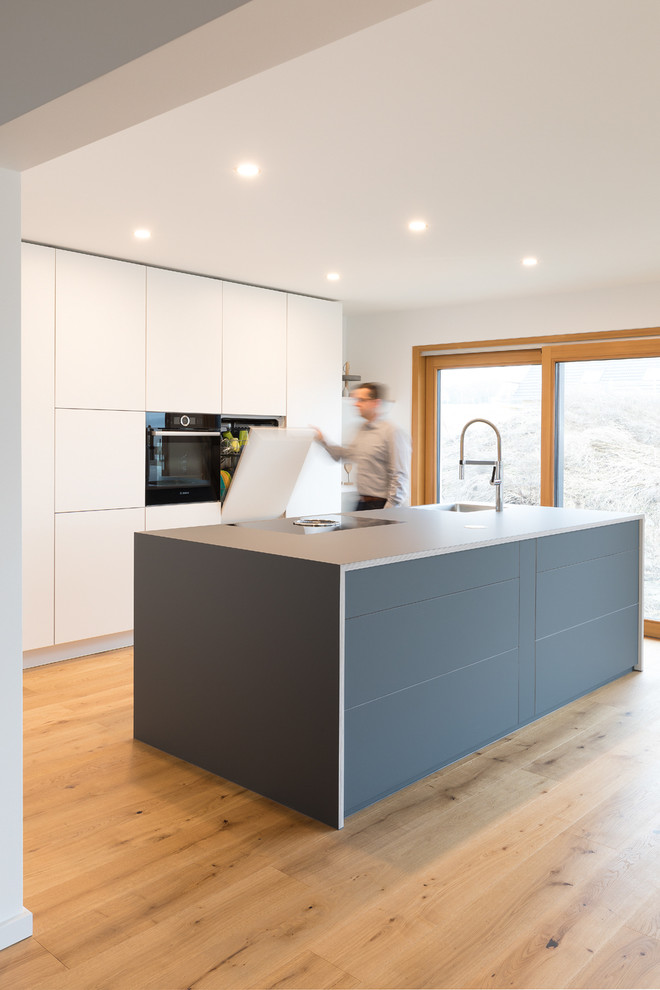 Design ideas for a large modern single-wall separate kitchen in Stuttgart with flat-panel cabinets, laminate benchtops, black appliances, with island, a single-bowl sink, white cabinets, white splashback, timber splashback, light hardwood floors, brown floor and black benchtop.