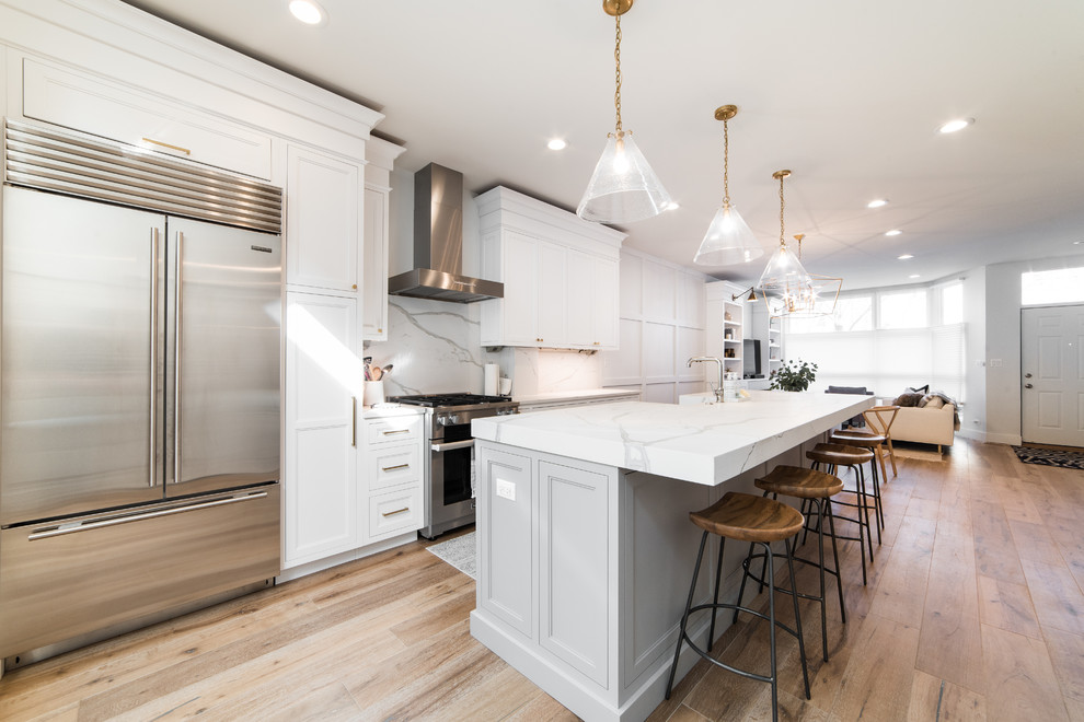 Inspiration for a mid-sized transitional galley open plan kitchen in Chicago with a farmhouse sink, recessed-panel cabinets, white cabinets, quartz benchtops, stone slab splashback, stainless steel appliances, light hardwood floors, with island and beige floor.