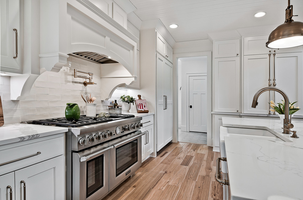Large arts and crafts open plan kitchen in Little Rock with a farmhouse sink, raised-panel cabinets, white cabinets, quartz benchtops, white splashback, stone tile splashback, stainless steel appliances, light hardwood floors, with island, brown floor and white benchtop.