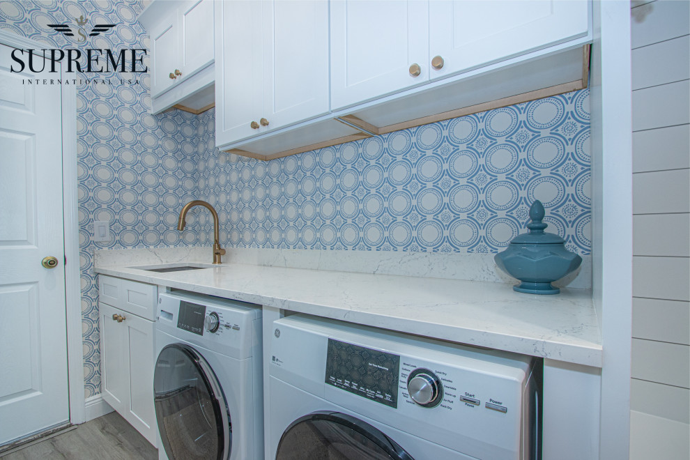 Large transitional single-wall dedicated laundry room in Tampa with an undermount sink, shaker cabinets, white cabinets, quartz benchtops, blue walls, porcelain floors, a side-by-side washer and dryer, brown floor, white benchtop, vaulted and wallpaper.