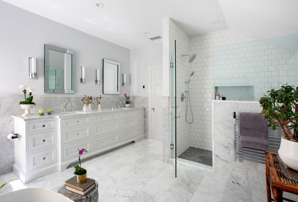 Photo of a mid-sized transitional master bathroom in New York with recessed-panel cabinets, white cabinets, a freestanding tub, an alcove shower, marble, grey walls, marble floors, an undermount sink, marble benchtops, white floor, a hinged shower door, white benchtops, a double vanity, a built-in vanity, decorative wall panelling, white tile and a niche.