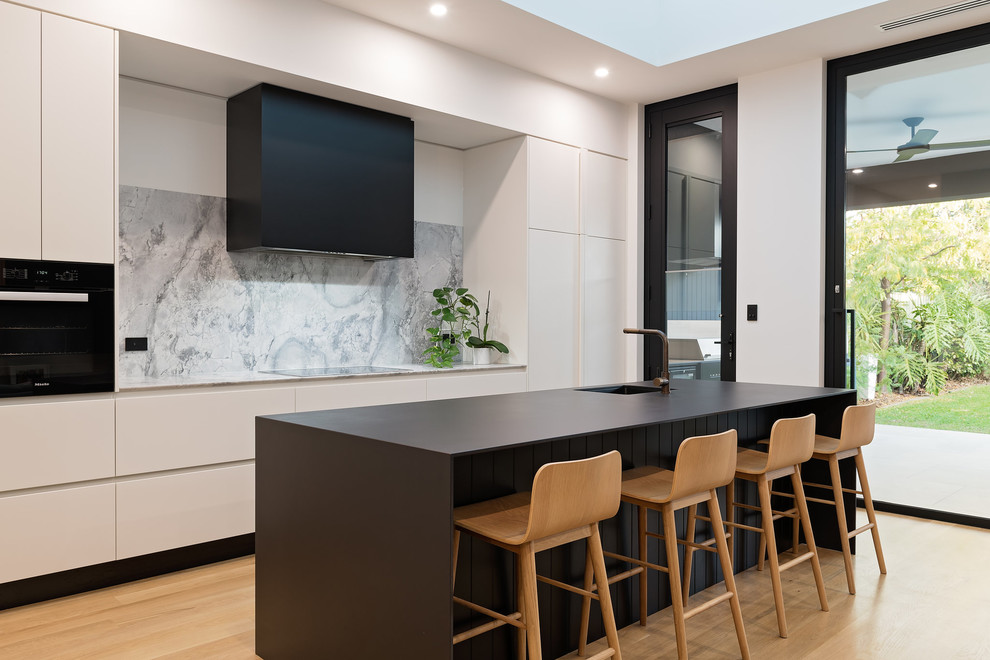 Photo of a contemporary galley kitchen in Adelaide with an integrated sink, flat-panel cabinets, grey splashback, black appliances, light hardwood floors, with island and black benchtop.