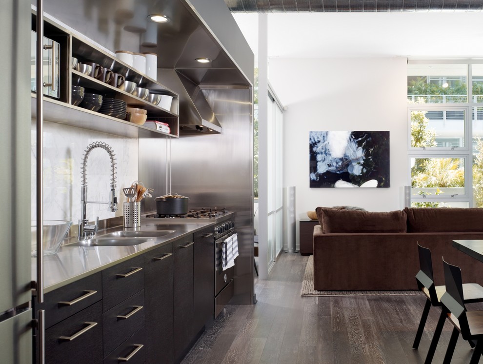 Photo of an industrial single-wall open plan kitchen in Los Angeles with stainless steel appliances, stainless steel benchtops, an integrated sink, open cabinets, white splashback, stone slab splashback, stainless steel cabinets and brown floor.