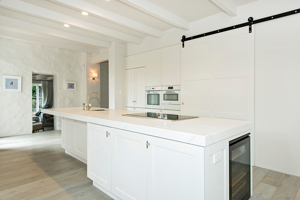 Photo of a large traditional l-shaped open plan kitchen in Auckland with an undermount sink, shaker cabinets, white cabinets, quartz benchtops, white splashback, white appliances, light hardwood floors, with island and beige floor.