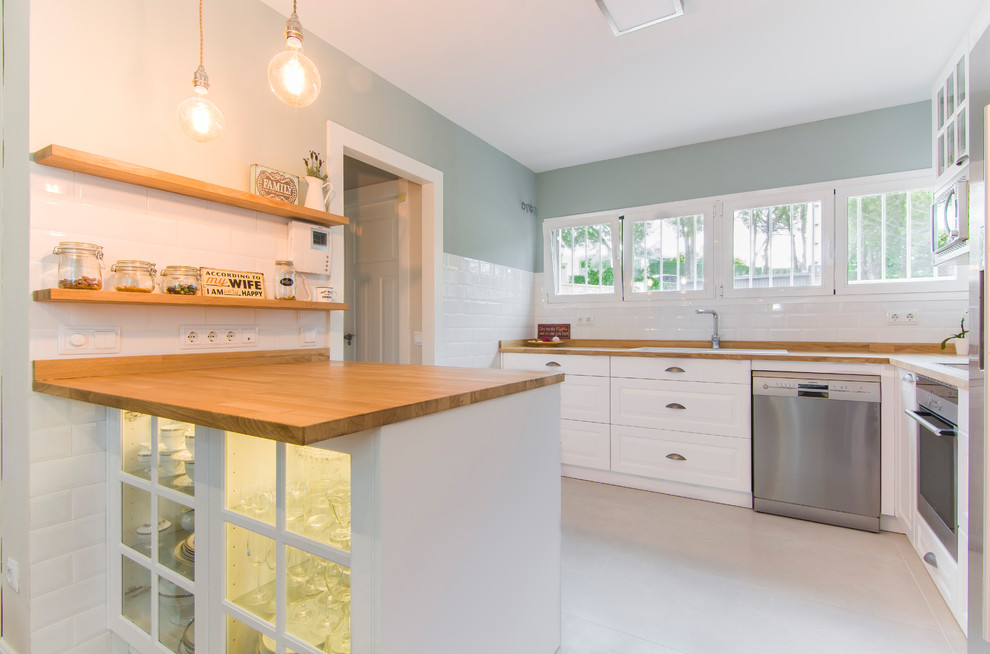 This is an example of a transitional u-shaped kitchen in Madrid with white cabinets, wood benchtops, stainless steel appliances, a peninsula, a drop-in sink, glass-front cabinets, white splashback, subway tile splashback, white floor and beige benchtop.