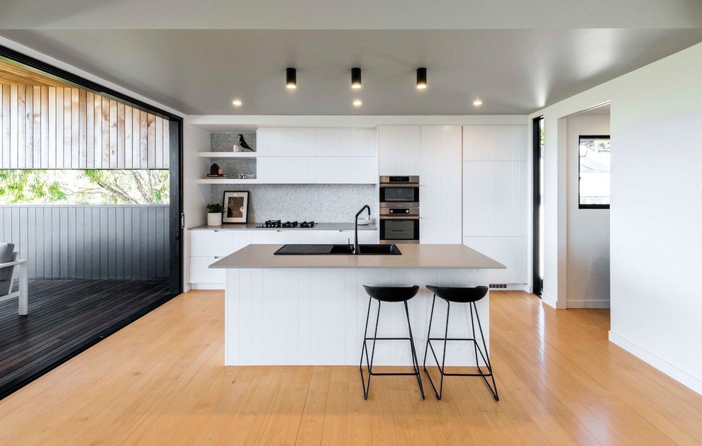 This is an example of a contemporary galley eat-in kitchen in Brisbane with a drop-in sink, grey splashback, light hardwood floors, with island, grey benchtop, flat-panel cabinets, white cabinets, mosaic tile splashback, panelled appliances and beige floor.