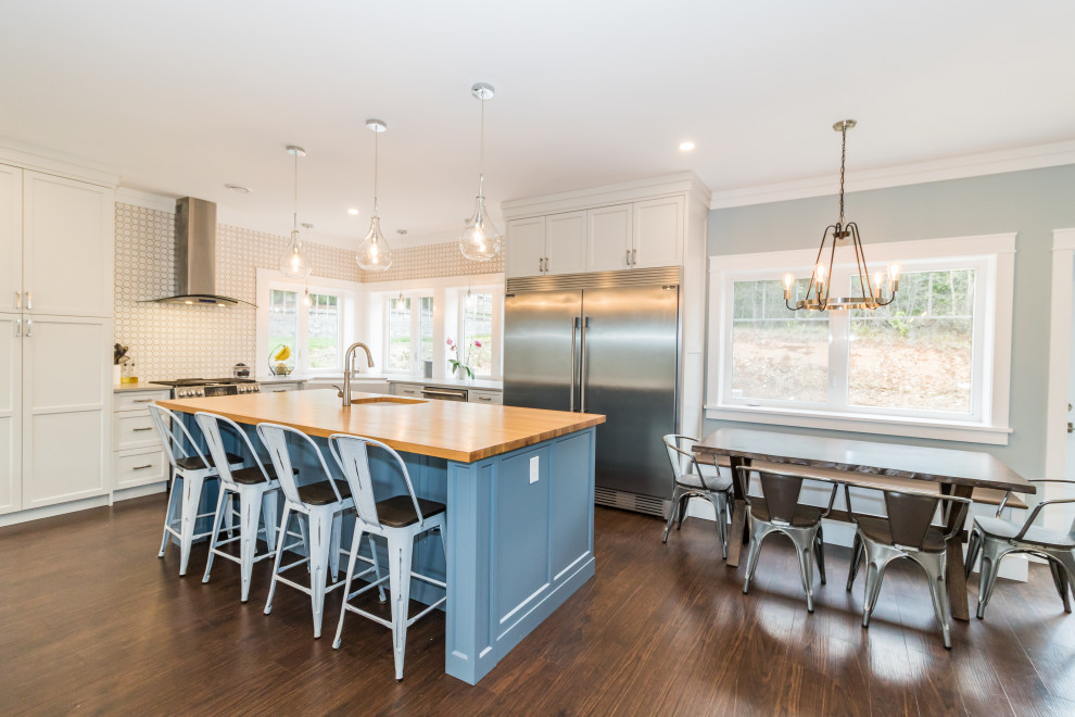 Mid-sized l-shaped vinyl floor and brown floor eat-in kitchen photo with a farmhouse sink, recessed-panel cabinets, white cabinets, wood countertops, multicolored backsplash, ceramic backsplash, stainless steel appliances and an island