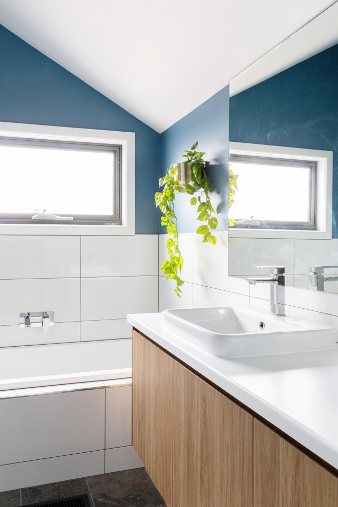 This is an example of a contemporary bathroom in Melbourne with flat-panel cabinets, medium wood cabinets, an alcove tub, white tile, blue walls, a drop-in sink, black floor, white benchtops, a floating vanity and vaulted.