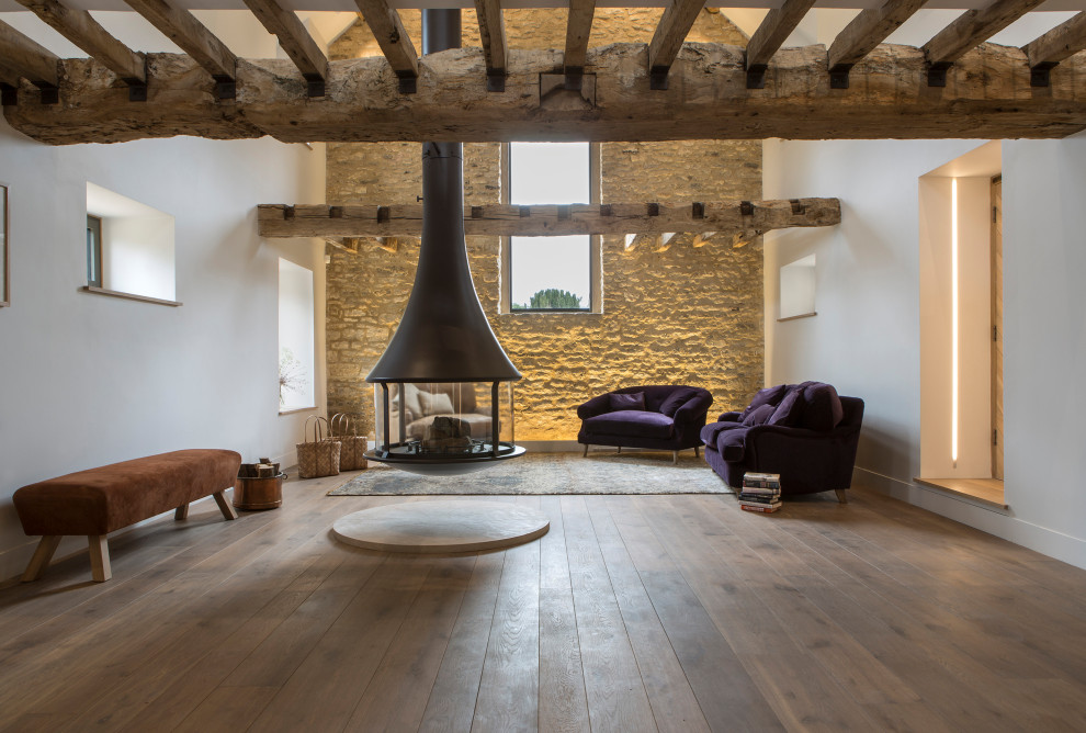 This is an example of a large country open concept living room in Oxfordshire with white walls, medium hardwood floors, a hanging fireplace and exposed beam.