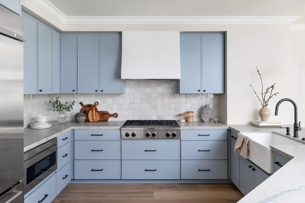 This is an example of a large transitional kitchen in San Diego with a farmhouse sink, blue cabinets, grey splashback, stainless steel appliances, light hardwood floors, a peninsula, brown floor, grey benchtop, beaded inset cabinets, quartzite benchtops and terra-cotta splashback.