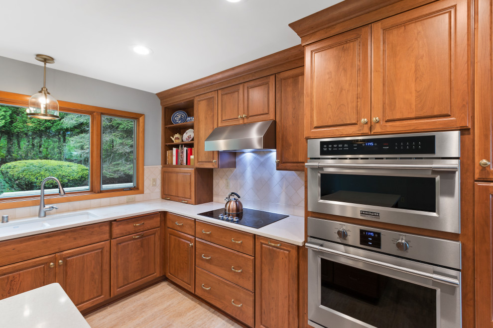 Design ideas for a mid-sized traditional l-shaped eat-in kitchen in Milwaukee with an undermount sink, raised-panel cabinets, brown cabinets, quartz benchtops, beige splashback, stone tile splashback, stainless steel appliances, vinyl floors, with island, brown floor and beige benchtop.