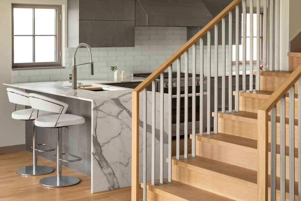 This is an example of a beach style l-shaped open plan kitchen in Portland Maine with a single-bowl sink, flat-panel cabinets, grey cabinets, marble benchtops, white splashback, glass tile splashback, stainless steel appliances, medium hardwood floors, a peninsula, beige floor and white benchtop.