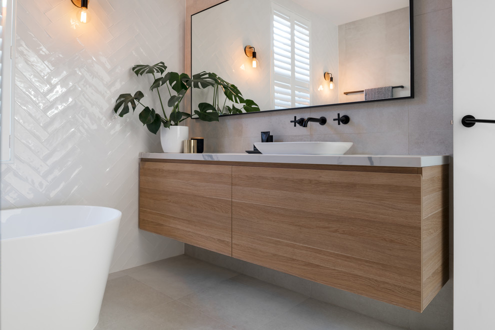 Inspiration for a mid-sized contemporary kids bathroom in Adelaide with flat-panel cabinets, light wood cabinets, porcelain floors, a vessel sink, grey floor, a freestanding tub, a wall-mount toilet, white tile, porcelain tile, white walls, solid surface benchtops and an open shower.