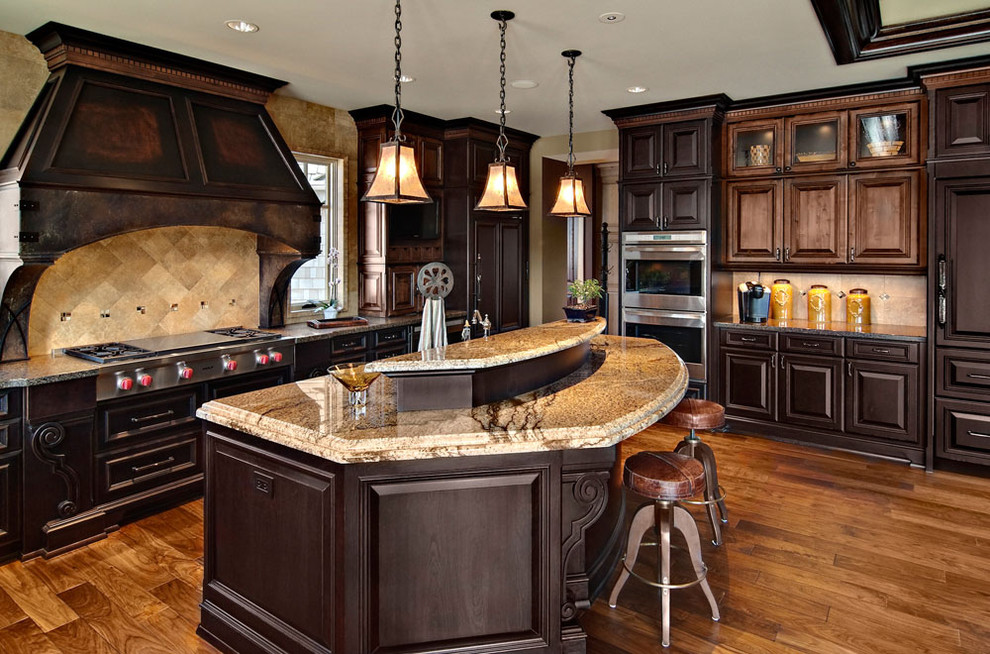 Traditional l-shaped open plan kitchen in Minneapolis with stainless steel appliances, raised-panel cabinets, dark wood cabinets, granite benchtops, brown splashback, stone tile splashback, medium hardwood floors and with island.