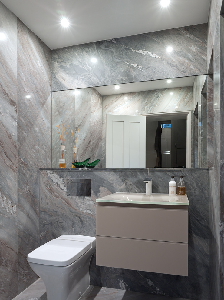 Photo of a mid-sized contemporary powder room in Kent with flat-panel cabinets, a one-piece toilet, beige tile, stone slab, beige walls, marble floors, a drop-in sink and glass benchtops.