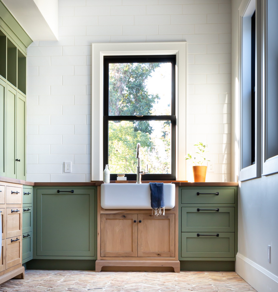 Design ideas for a large farmhouse l-shaped separated utility room in San Francisco with a belfast sink, shaker cabinets, green cabinets, wood worktops, white splashback, porcelain splashback, white walls, brick flooring, an integrated washer and dryer, beige floors and brown worktops.
