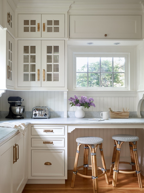 Cozy cottage kitchen with white shaker RTA cabinets, glass doors, brass hardware, beadboard backsplash, marble counters, and rattan stools by a window.