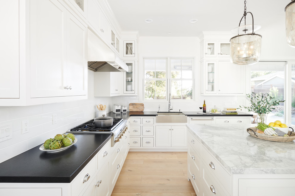 Inspiration for a country l-shaped open plan kitchen in San Francisco with a farmhouse sink, shaker cabinets, white cabinets, white splashback, subway tile splashback, stainless steel appliances, light hardwood floors, with island and black benchtop.