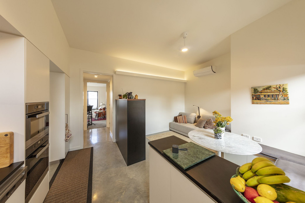 This is an example of a small contemporary galley eat-in kitchen in Canberra - Queanbeyan with flat-panel cabinets, white cabinets, grey splashback, ceramic splashback, stainless steel appliances, concrete floors, with island, grey floor and black benchtop.